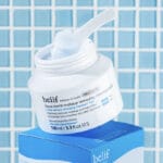 belif Cleansing Balm 100ml - Baume démaquillant hydratant à la fleur de lotus & vitamine E. Image avec applicateur, sans couvercle, carrelage de salle de bain en arrière-plan.