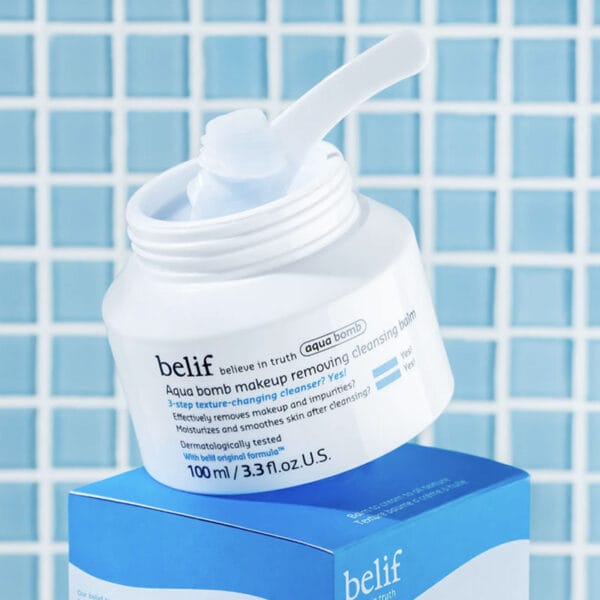 belif Cleansing Balm 100ml - Baume démaquillant hydratant à la fleur de lotus & vitamine E. Image avec applicateur, sans couvercle, carrelage de salle de bain en arrière-plan.