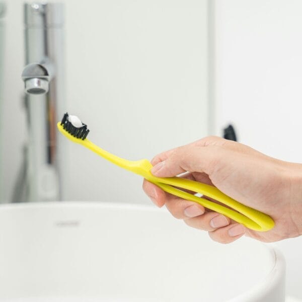 megasmile Loop Zahnbürste mit Aktivkohle-Borsten. Abbildung Zahnbürste yellow in der Hand, über dem Waschbecken.